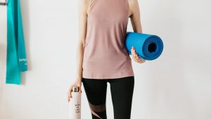 Faceless slim female athlete in sportswear standing with blue fitness mat and water bottle while preparing for indoors workout